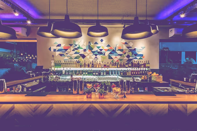 Modern bar interior with a wooden counter, colorful geometric wall art, hanging pendant lights, and shelves stocked with liquor bottles and glassware.