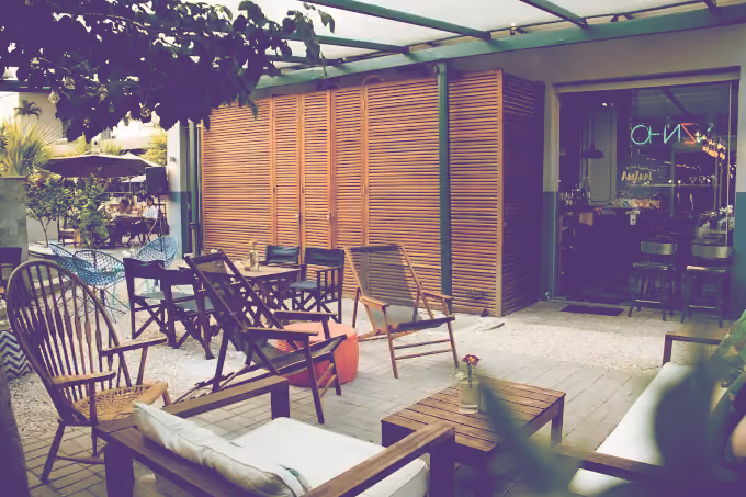 Outdoor seating area of a gastrobar with wooden chairs and tables, a shaded pergola, and greenery.