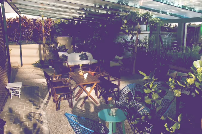 Cozy outdoor seating area with wooden chairs and tables under a glass roof surrounded by greenery.