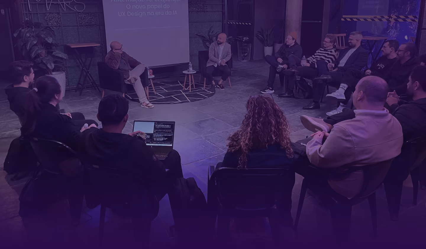 Group of people seated in a circle in a dimly lit room, listening to two speakers in the center with a laptop and a small projector screen displaying text in Portuguese.