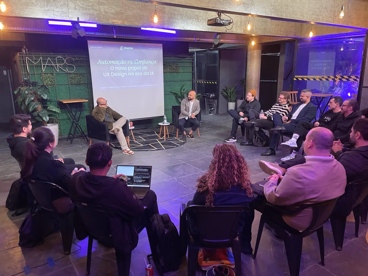 Small group of people seated in a circle in an indoor modern setting with industrial decor, listening to two speakers seated on chairs near a projector screen displaying a presentation titled 'Automação vs. Confiança: O novo papel do UX Design na era da IA'.
