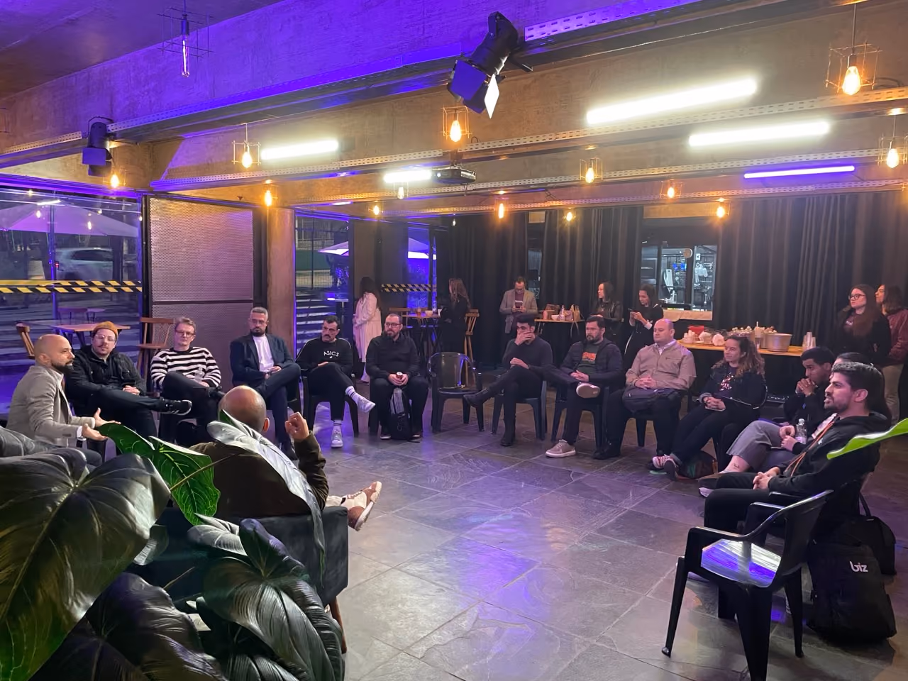 A group of people seated in a semi-circle in a modern indoor setting with purple and warm lighting, engaged in a discussion or meeting.