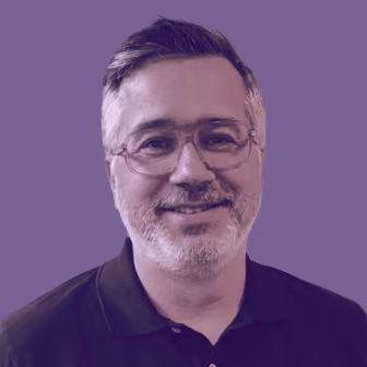 Middle-aged man with glasses, short gray hair, and a beard smiling, wearing a black collared shirt against a plain purple background.