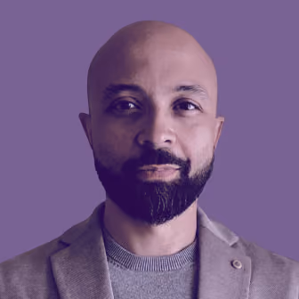 Portrait of a bald man with a beard and mustache wearing a grey blazer and sweater, against a purple background.