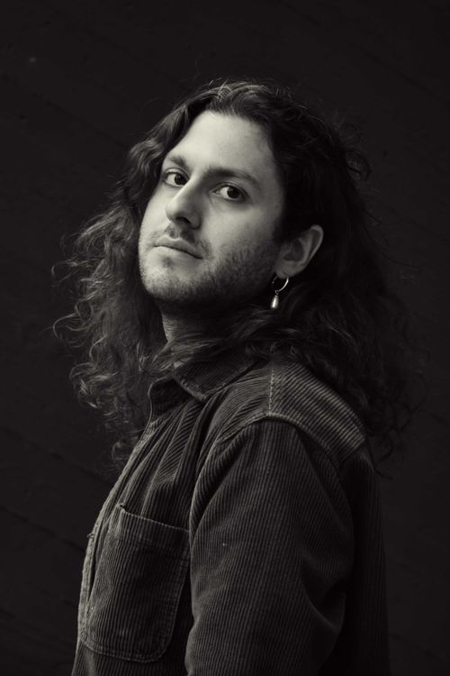Black and white portrait of a man with long curly hair and a hoop earring, gazing thoughtfully at the camera in a moody, artistic style.
