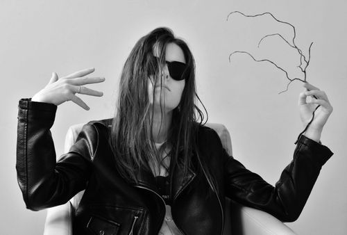 Artistic black and white portrait of a woman in sunglasses and a leather jacket holding a twisted branch, with hair covering part of her face in a bold, expressive pose.