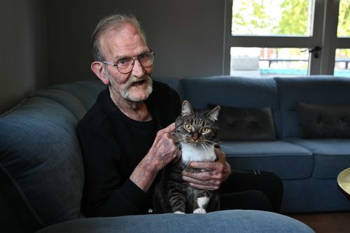 Elderly man with glasses and a mustache sitting on a couch while holding a tabby cat with white markings, both looking toward the camera in a cozy living room.