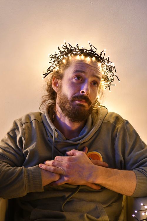 Bearded man with a contemplative expression wearing a hoodie, sitting with hands clasped on his chest and fairy lights arranged like a glowing halo around his head, creating a humorous and artistic visual effect.