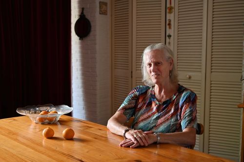 Elderly woman smiling while sitting at a wooden table with a bowl of oranges, warmly lit by natural light in a cozy home interior.
