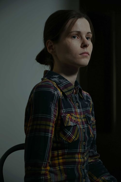 Moody portrait of a young woman (Polina Nasedkina) in a plaid shirt sitting in soft, directional lighting, gazing pensively into the distance.