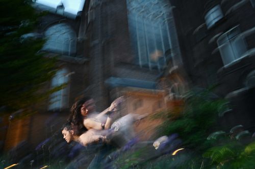 Artistic photo of two dancers in motion blur outside a large Gothic-style building, surrounded by flowers and dramatic twilight lighting.