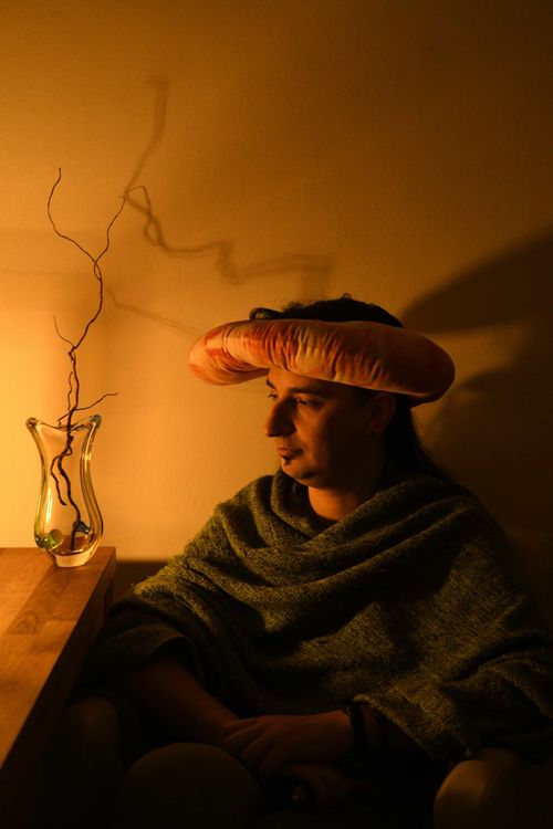 Person in dim, moody lighting wearing a plush sausage hat and green shawl, sitting beside a wooden table with a glass vase holding decorative twigs.