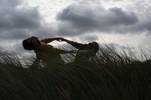 Two dancers perform an expressive movement sequence in tall coastal grass under a dramatic, cloudy sky, silhouetted by natural light.