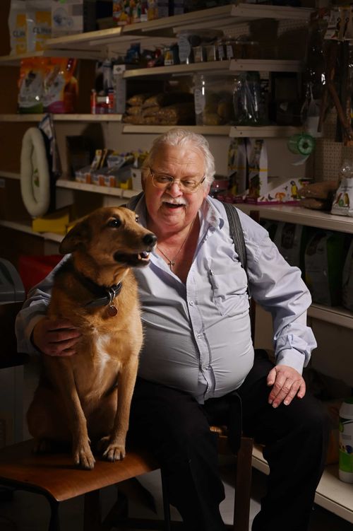 Smiling elderly man sitting with a brown dog on a bench inside a pet store, surrounded by pet food and supplies.