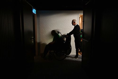 Elderly man in a wheelchair being pushed by another senior through a dimly lit hallway with a white brick wall and emergency exit sign overhead.