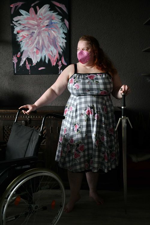 Woman wearing a floral dress and pink face mask stands barefoot indoors, holding a sword in one hand and a wheelchair in the other, with abstract pink artwork on the wall behind her.