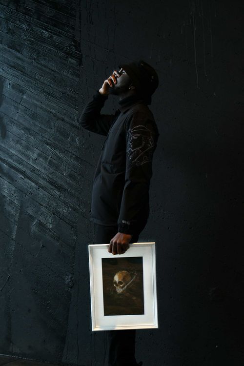 Man dressed in black, wearing sunglasses and a bucket hat, stands against a dark textured wall while talking on the phone and holding framed artwork featuring a skull.