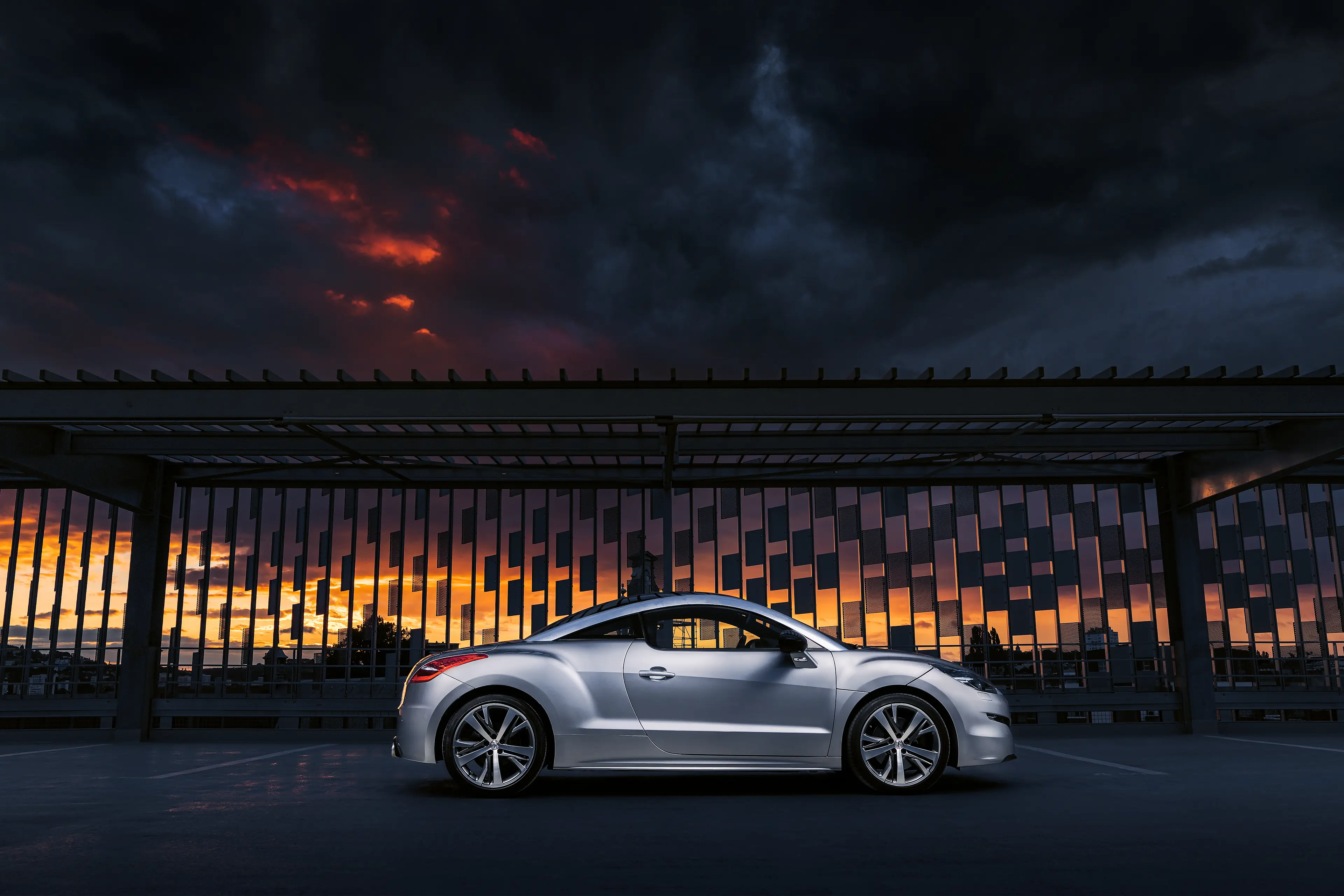 Peugeot RCZ grise de profil dans un parking sous un ciel menaçant au coucher du soleil au flash