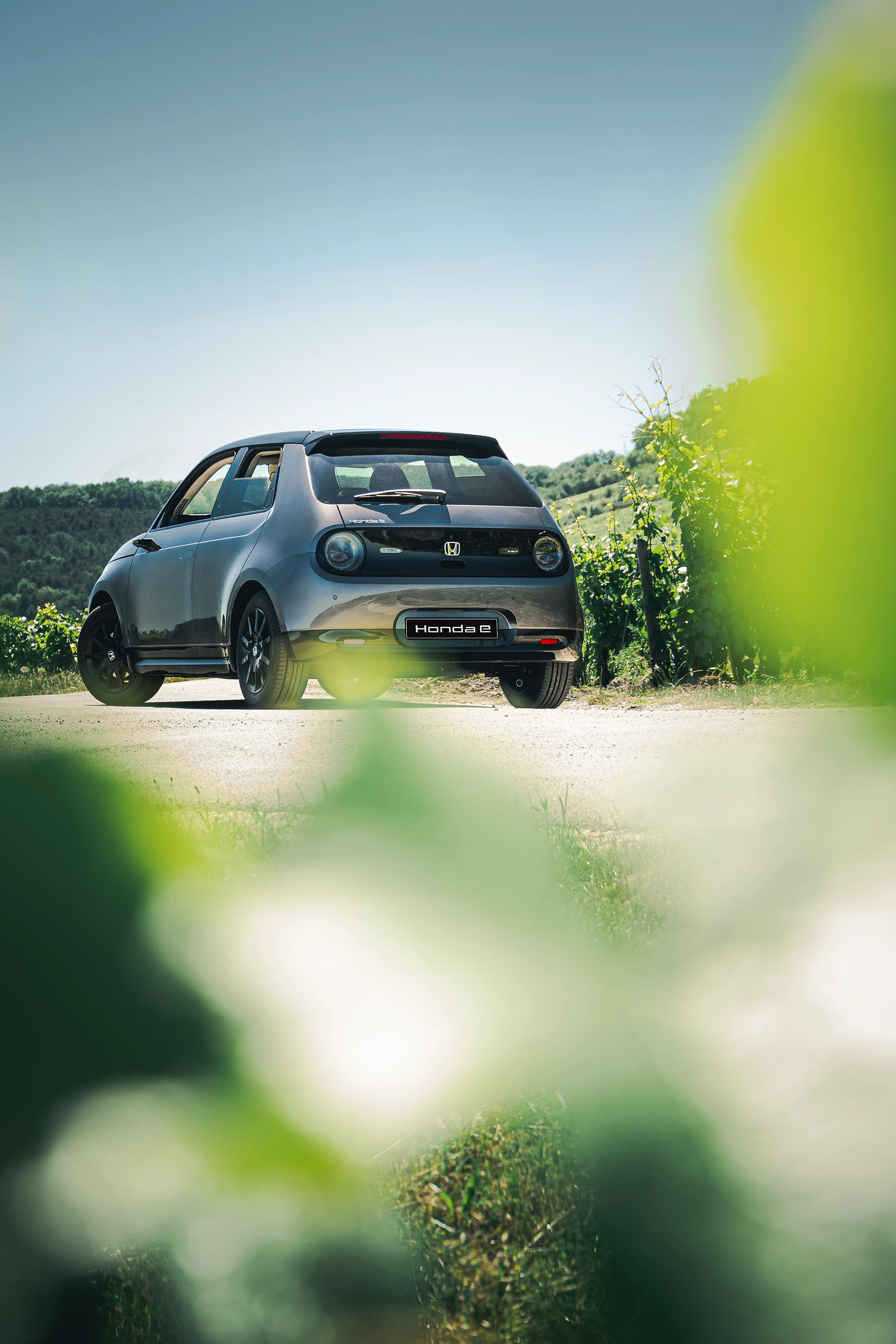 Honda E grise anthracite de 3/4 arrière avec des feuilles de vigne floues au premier plan
