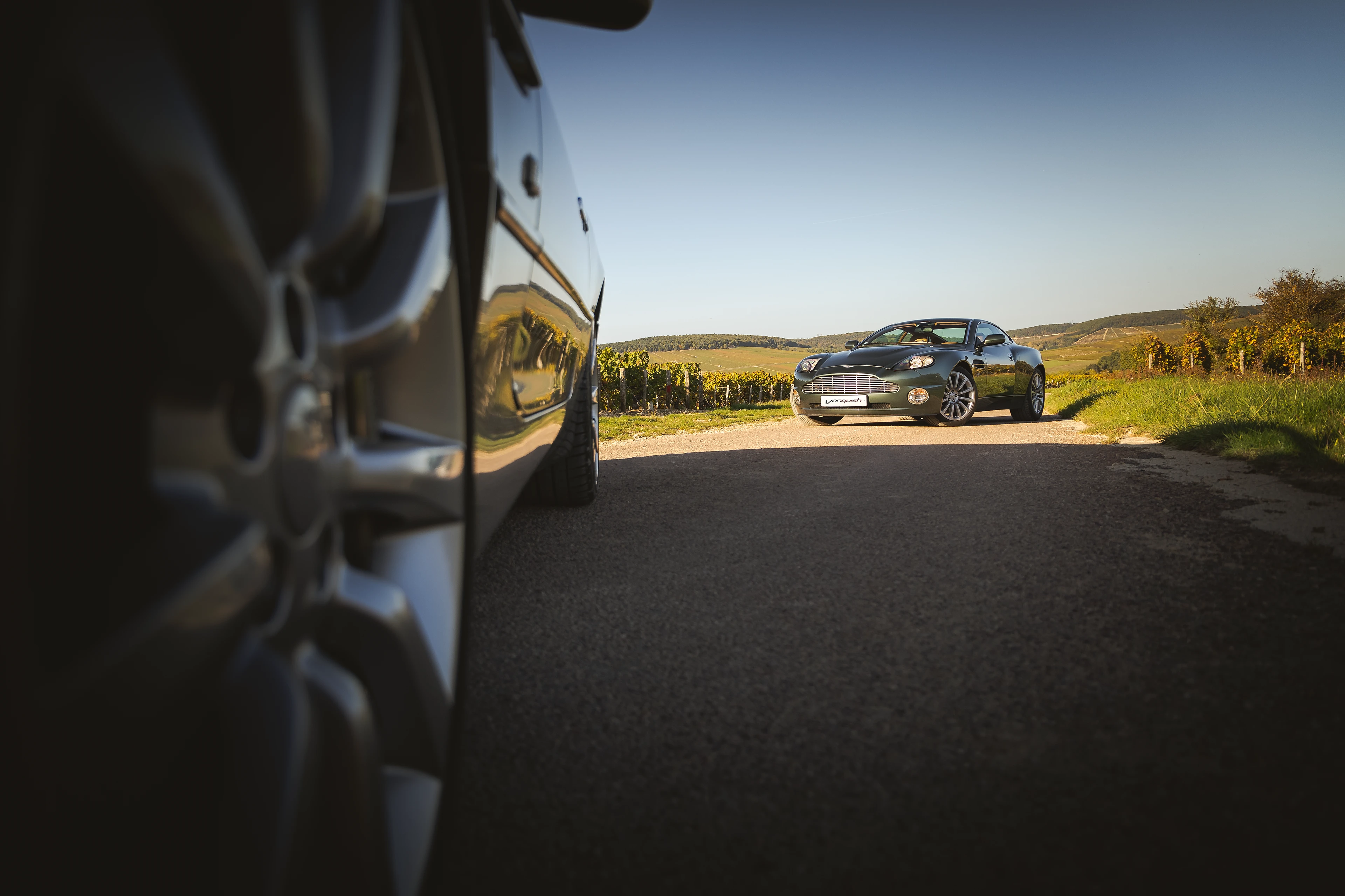 Aston Martin Vanquish verte de 3/4 avant avec une jante d'Aston Martin DB7 floue au premier plan