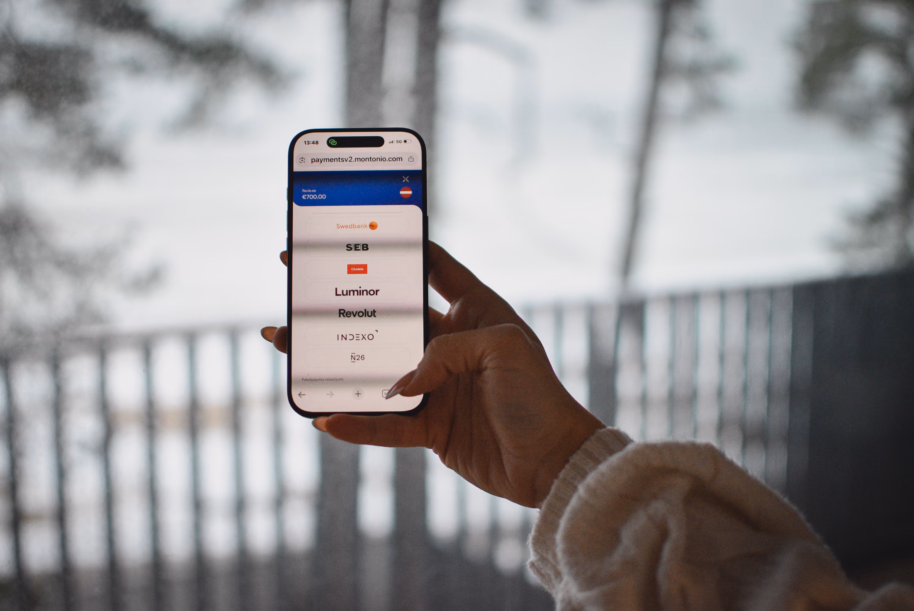 A close-up view of a hand holding a smartphone, displaying the Montonio payment gateway on the screen. The payment interface shows a total of €700.00 and a vertical list of bank selection options, including Swedbank, SEB, Luminor, and Revolut. The person holding the phone is wearing a chunky, cream-colored knit sweater. The background is a heavily blurred, cool-toned outdoor winter scene featuring a balcony railing and trees.