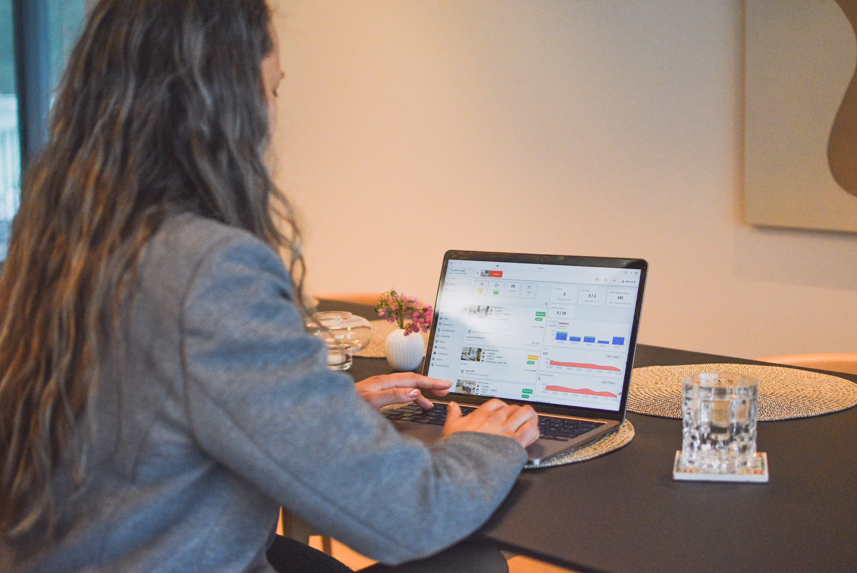 A person with long wavy hair wearing a gray blazer sits at a dark table, typing on a laptop. The laptop screen displays the BOUK software dashboard, showing various data charts, metrics, and property listings. On the table next to the laptop are a textured glass of water on a coaster, a round woven placemat, and a small white vase with pink flowers.