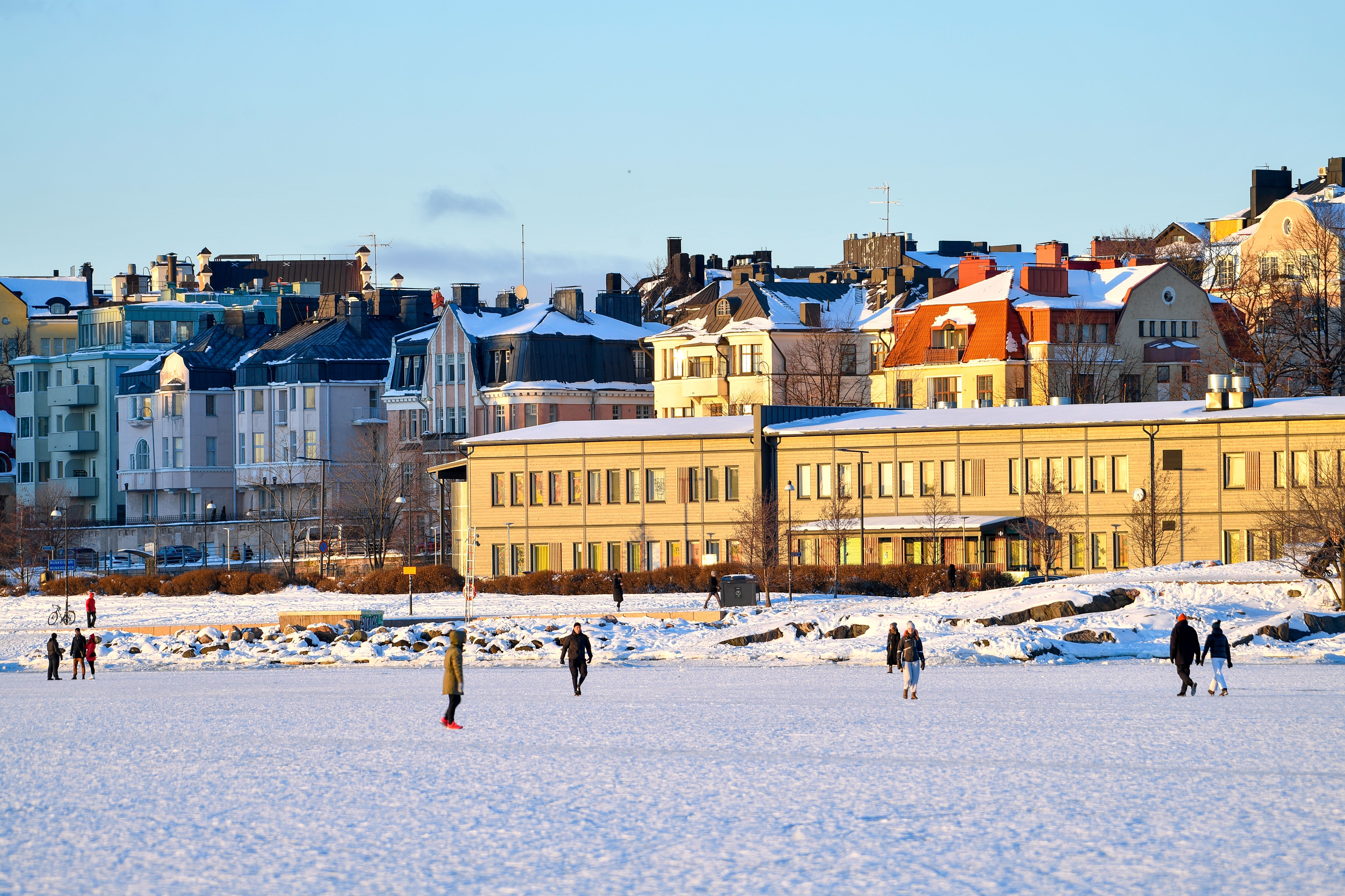 A serene winter scene of people walking on a frozen sea beside colorful Helsinki buildings. – Margo Evardson