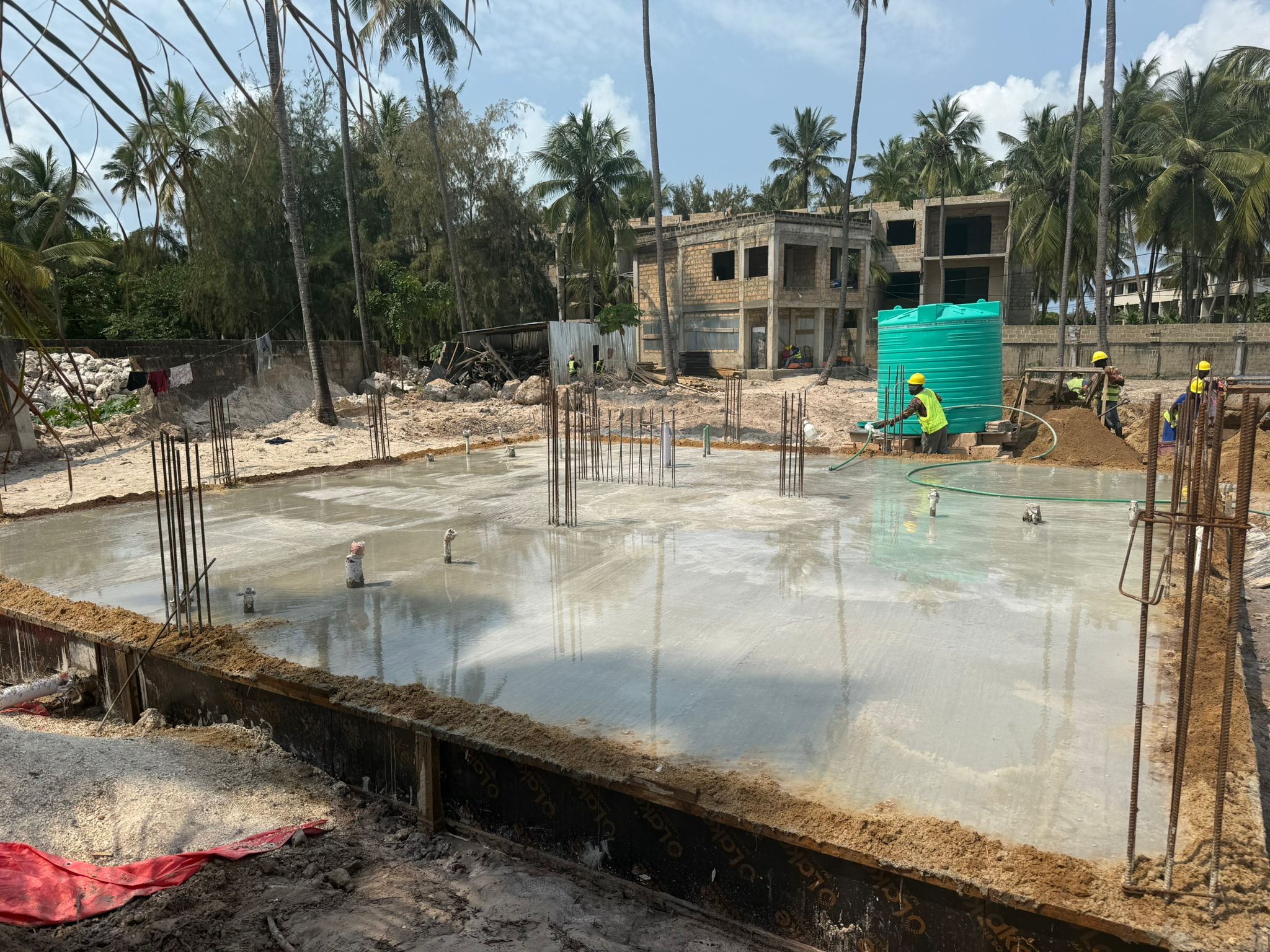 Workers smoothing freshly poured concrete for a building foundation in a tropical setting.