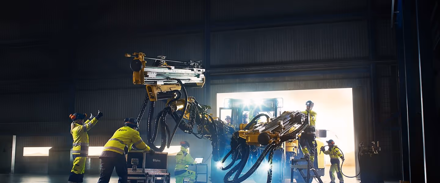 Arbeidere i verneutstyr opererer med en stor gul borerobot i et industrielt lagerbygg.