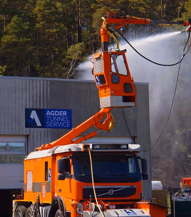 Oransje Volvo lastebil med bomløfteutstyr sprøyter vann nær en bygning med skiltet Agder Tunnel Service.