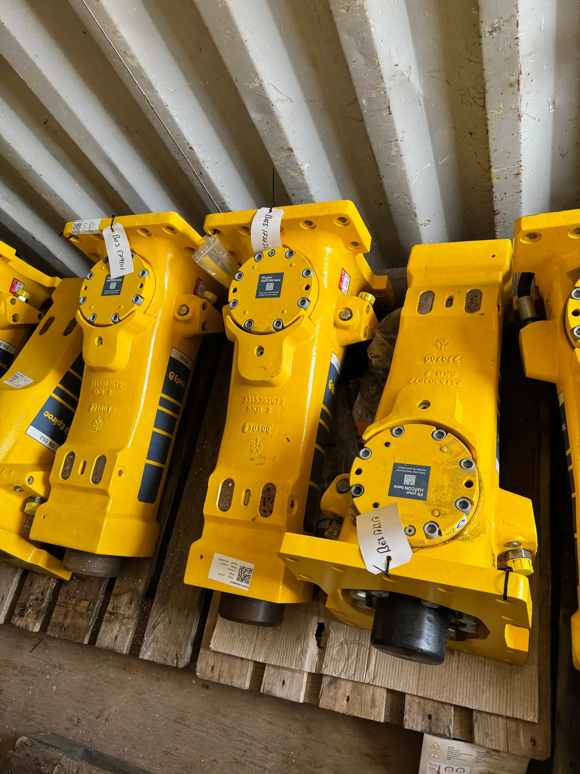 Flere gule hydrauliske maskindeler merket med etiketter og QR-koder, plassert på et trebord under et metalltak.