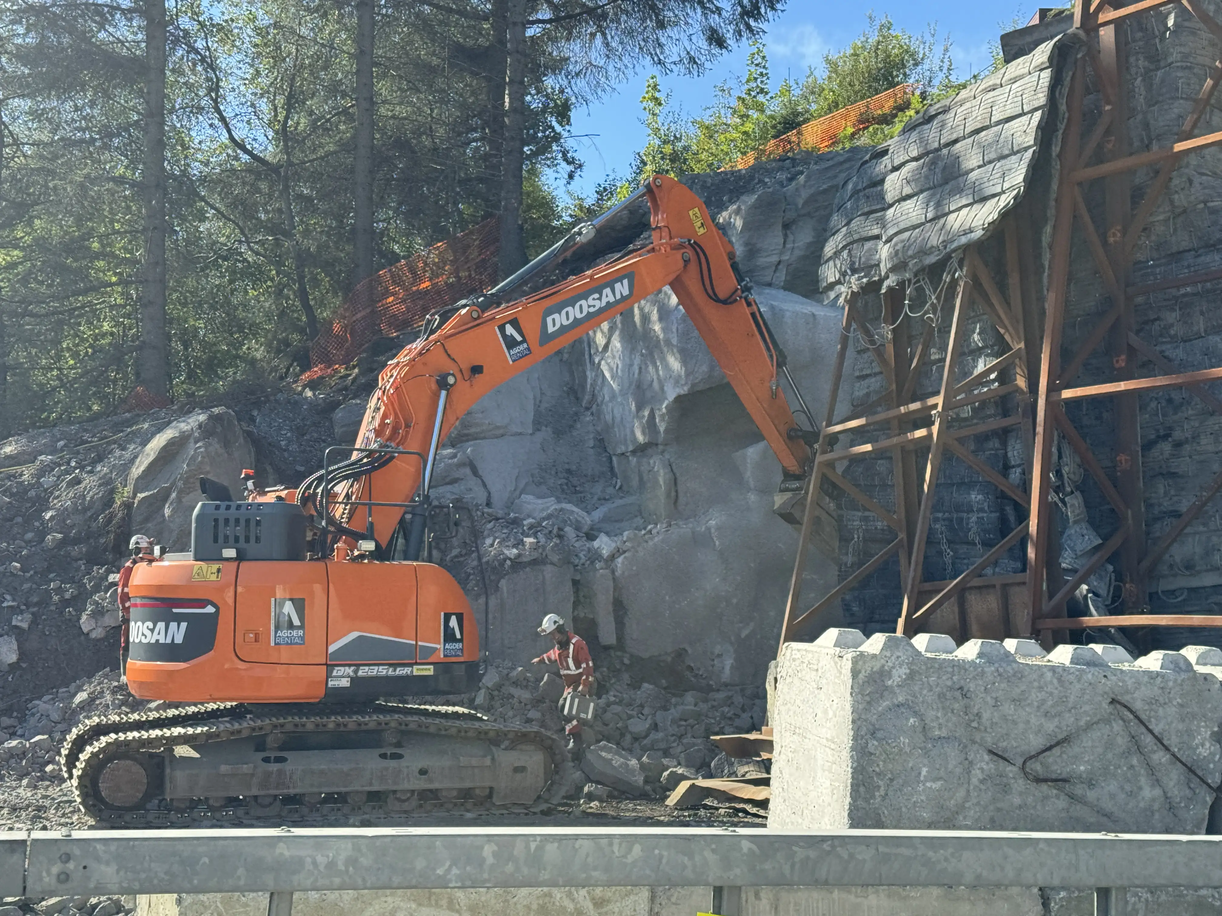 Oransje Doosan gravemaskin i arbeid nær en steinvegg med en bygningsarbeider i vernedrakt og hjelm ved siden av.