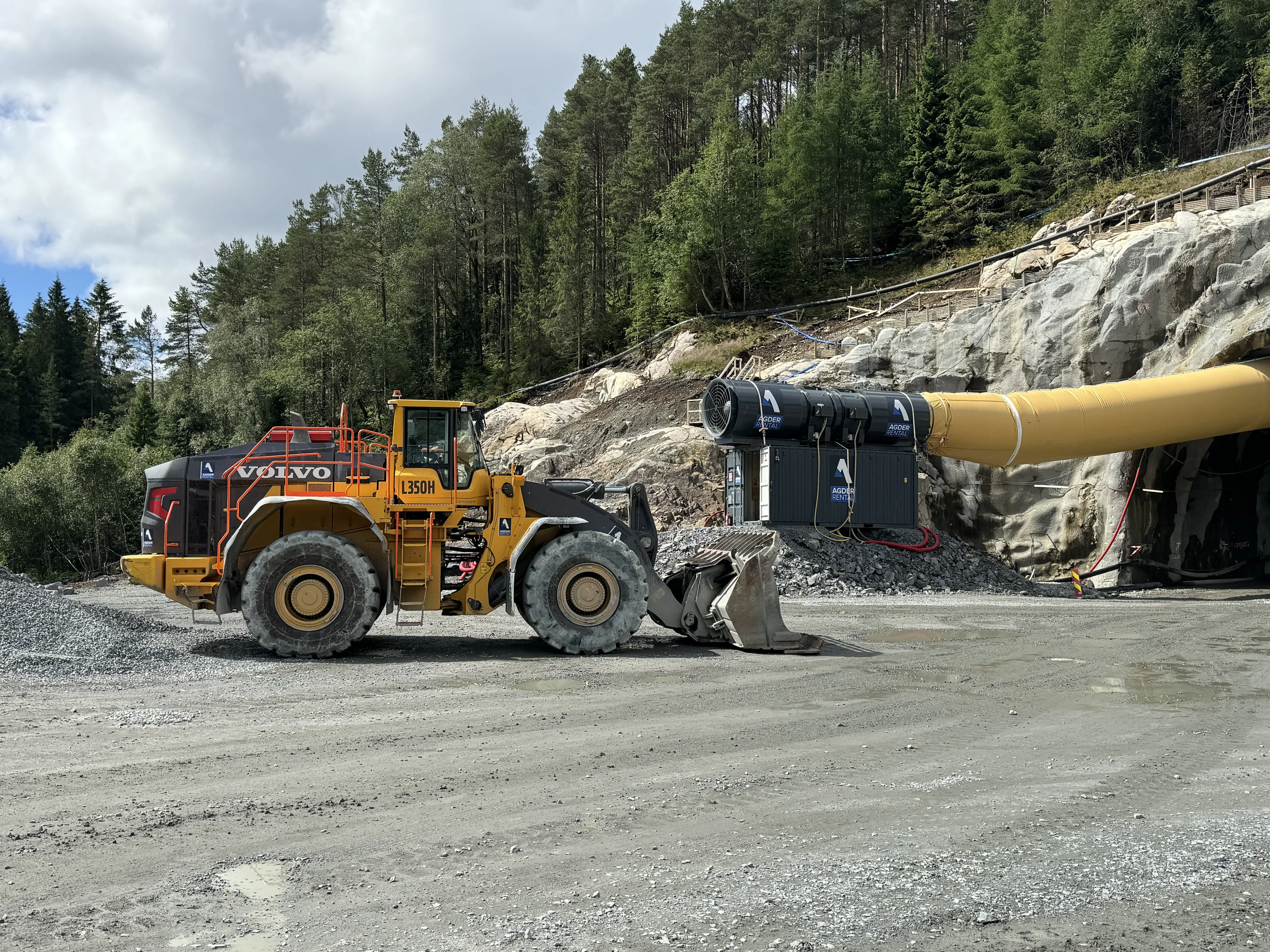 Gul Volvo L350H hjullaster ved inngangen til en tunnel i et skogsområde med rør og maskineri.