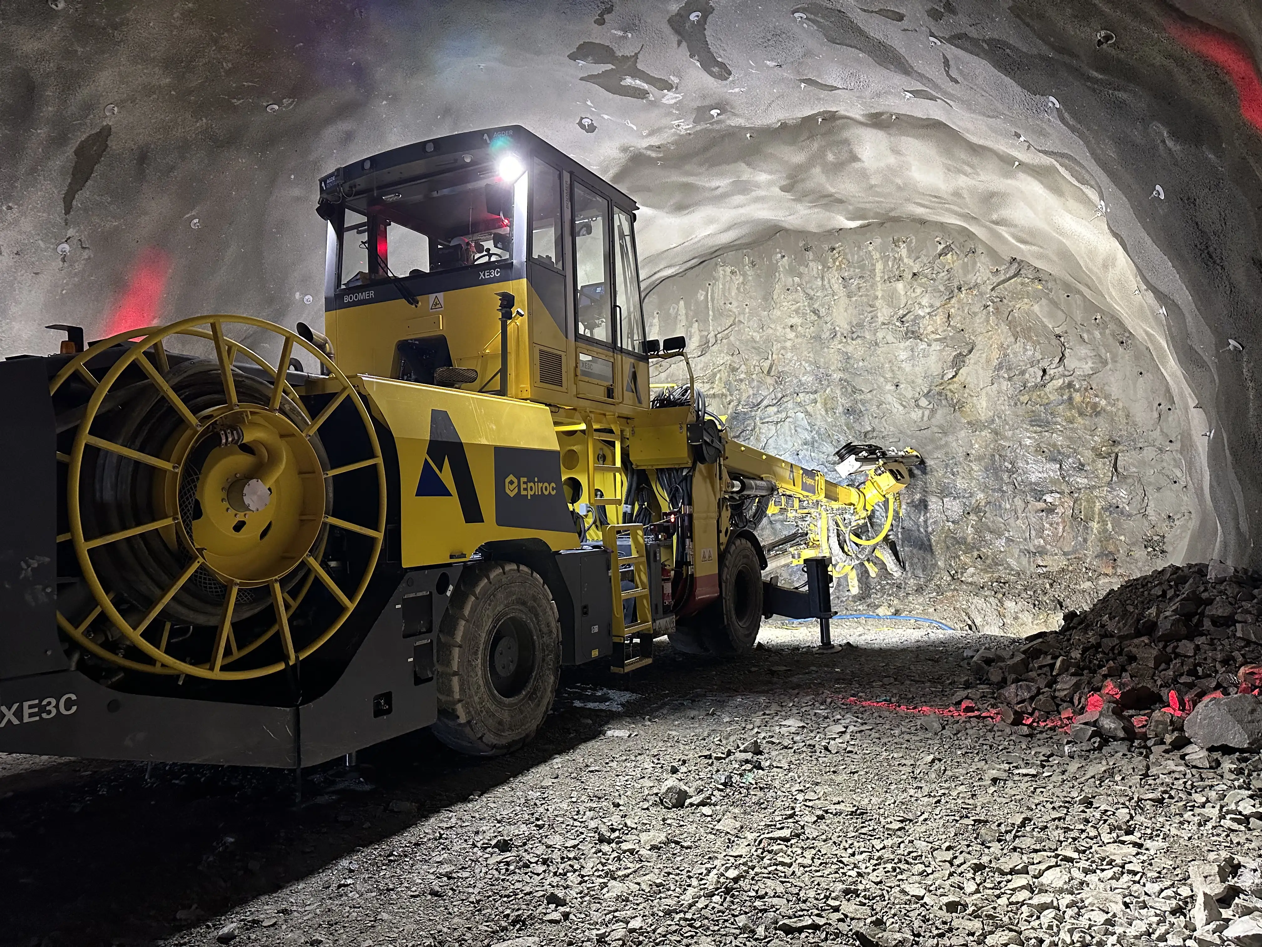 Gul Epiroc-gruvedriftsmaskin i tunnel på grusholdig underlag med fjellvegg belyst foran.