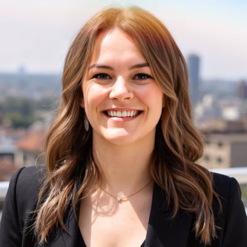 Professional headshot of Kelsea Gust, smiling woman with light brown hair, wearing a black jacket, with a city in the background.