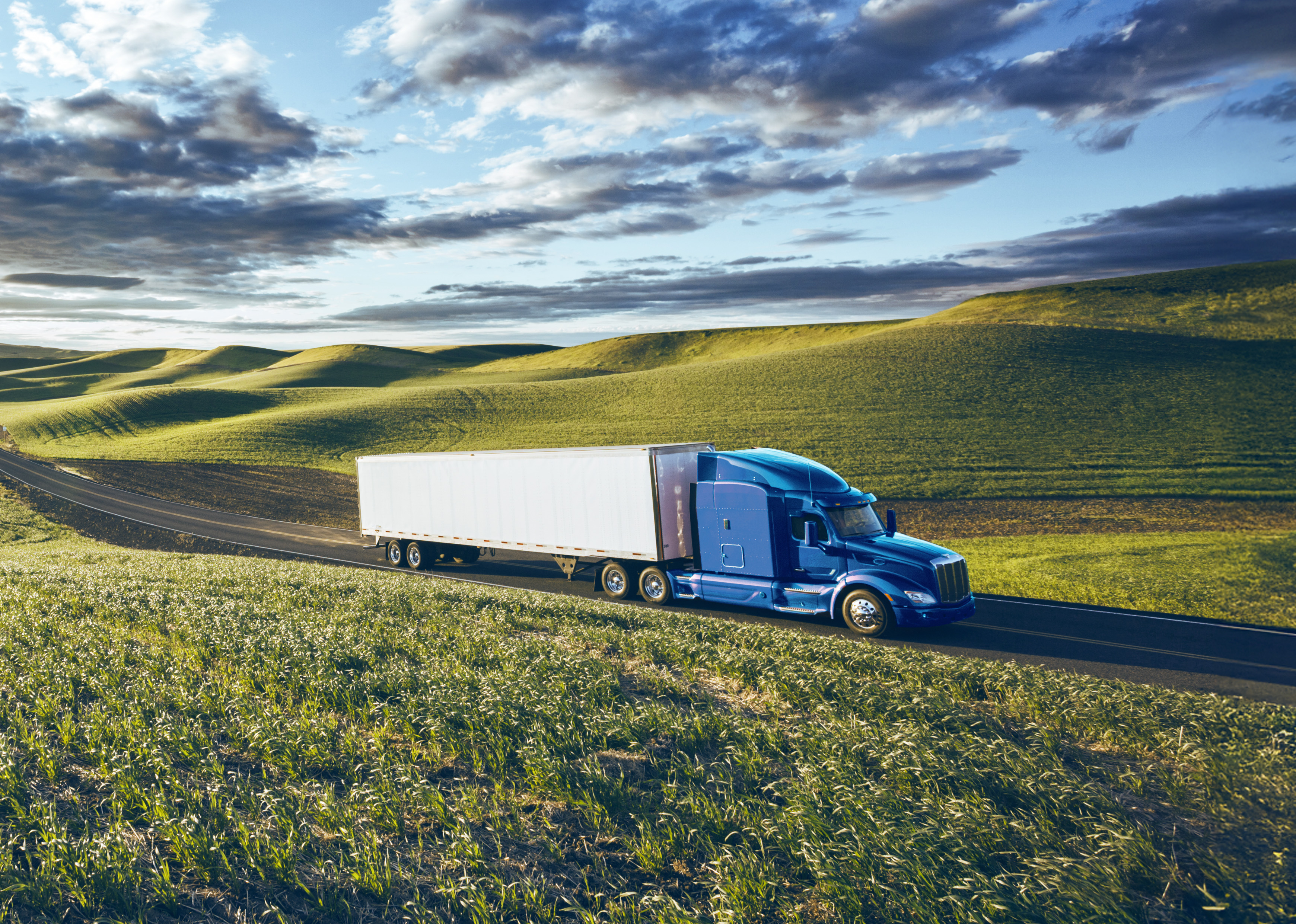 Camion semi-remorque bleu roulant sur une route bordée de collines vertes sous un ciel partiellement nuageux.