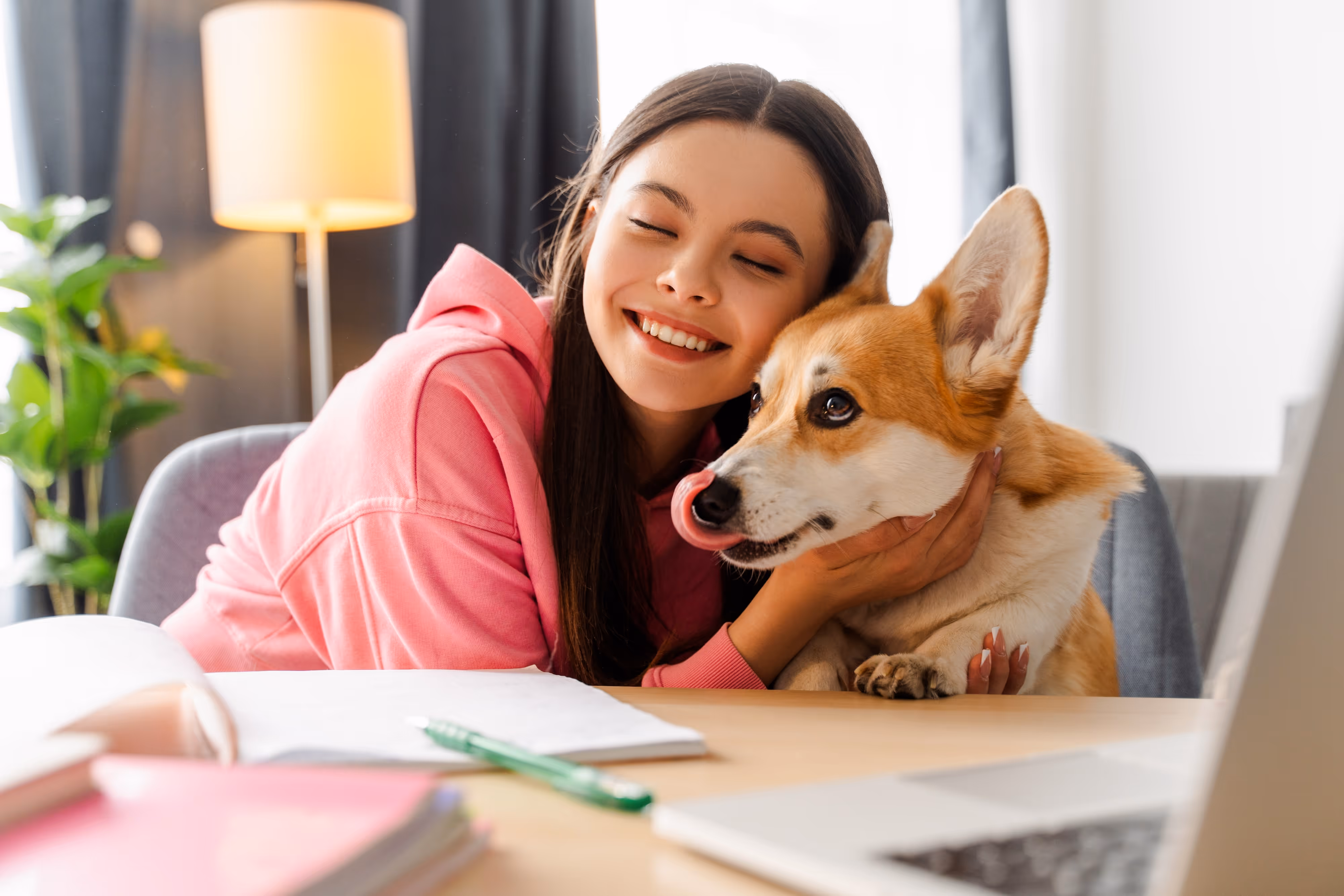 Smiling young student at a desk with a laptop, hugging a corgi dog, representing the pet-friendly and comfortable study environment at Global College Station in College Station, TX.