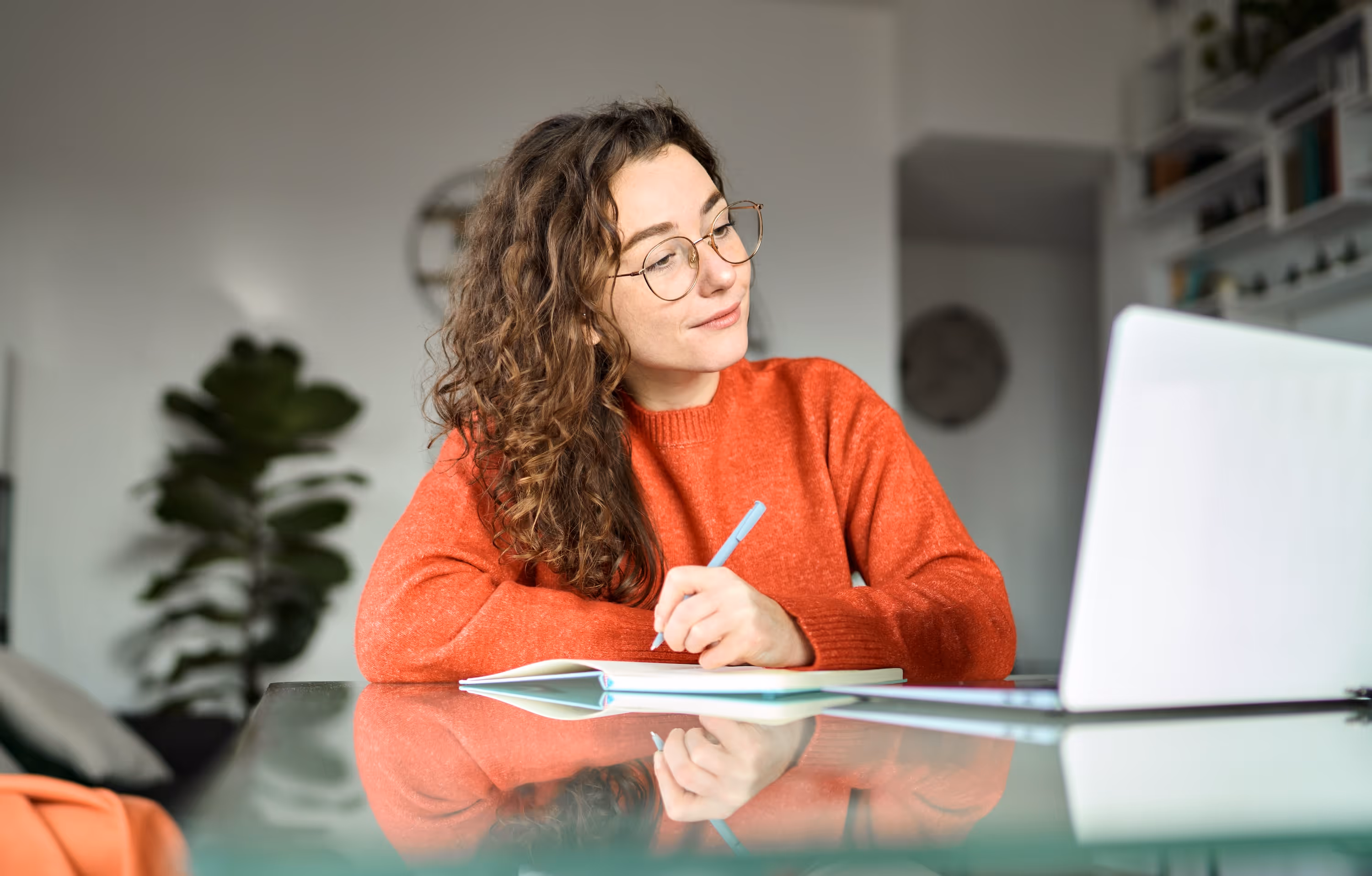 A focused female student wearing glasses and an orange sweater, studying and taking notes on her laptop in a bright apartment, representing the ideal academic environment at Global College Station in College Station, TX.
