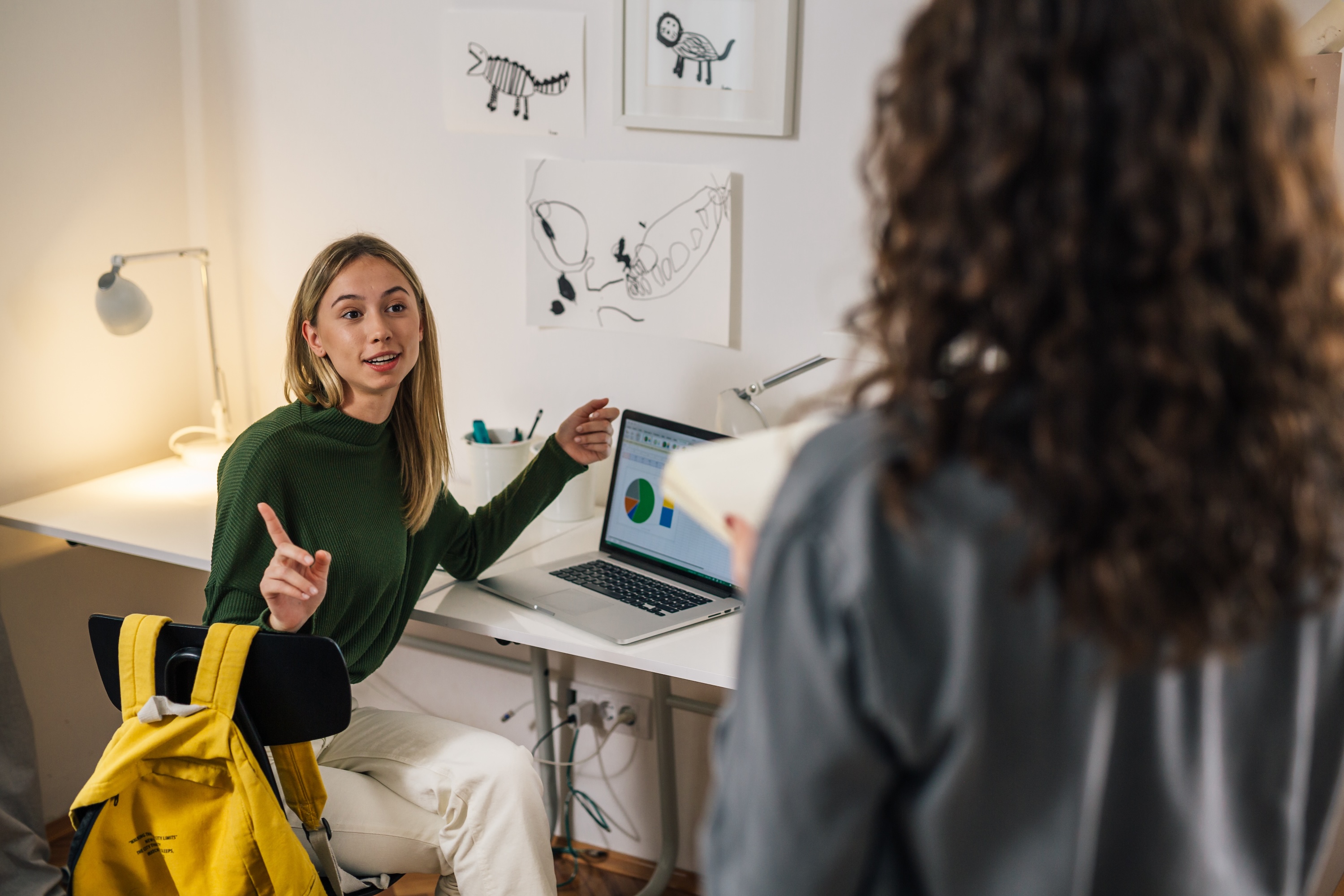A resident sits at a white desk with a laptop displaying charts, gesturing while speaking to another person in a bright, modern apartment office space at Global College Station.
