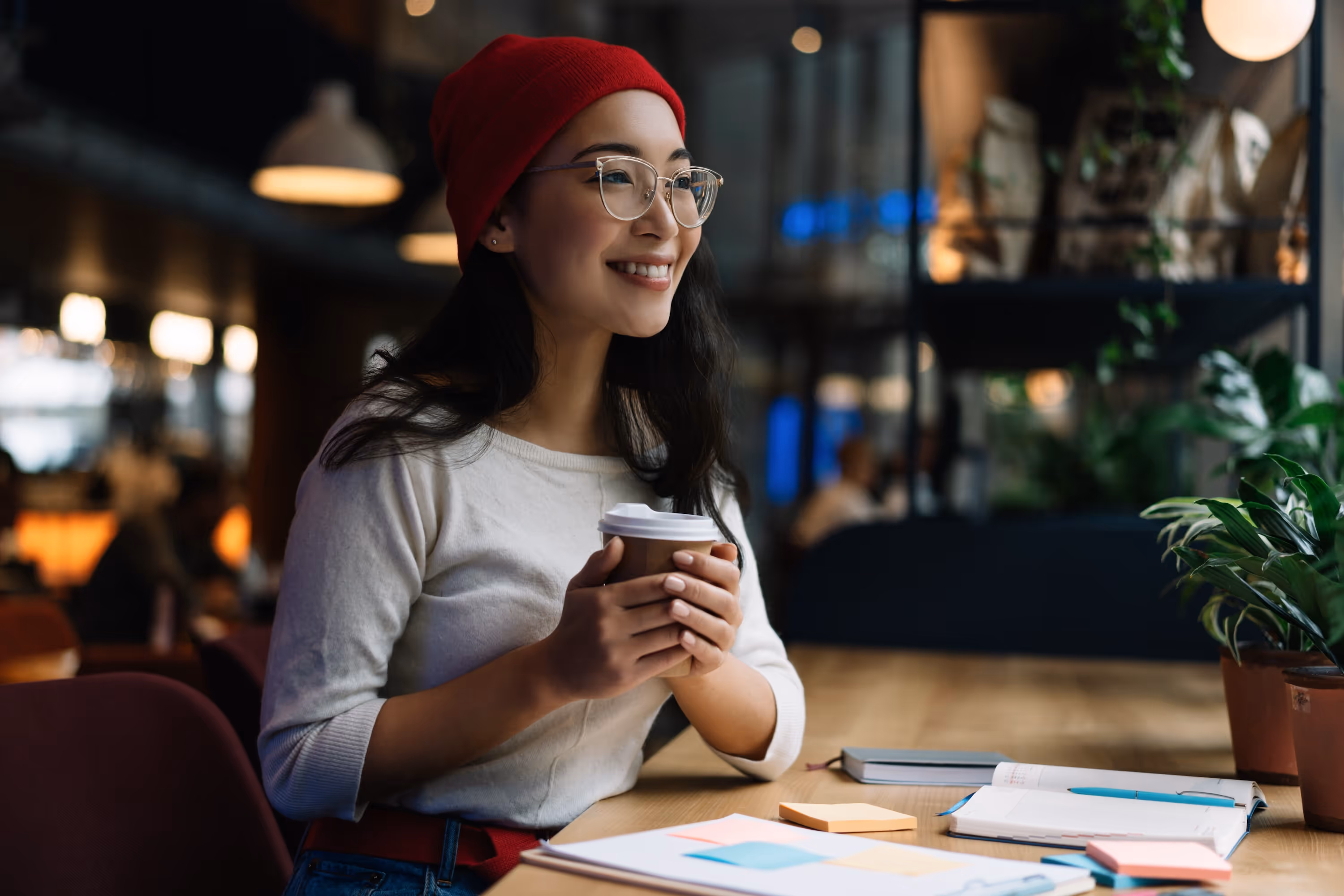 Student in a red beanie and glasses sitting at a cafe table in College Station with a coffee, notebook, and pens.