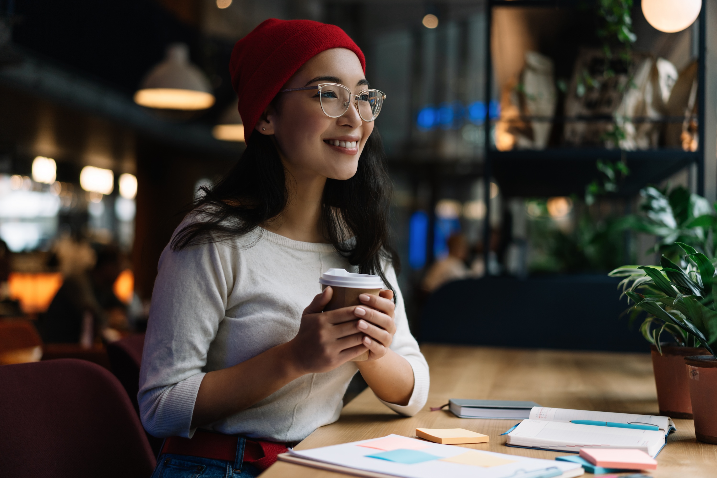 Student in a red beanie and glasses sitting at a cafe table in College Station with a coffee, notebook, and pens.