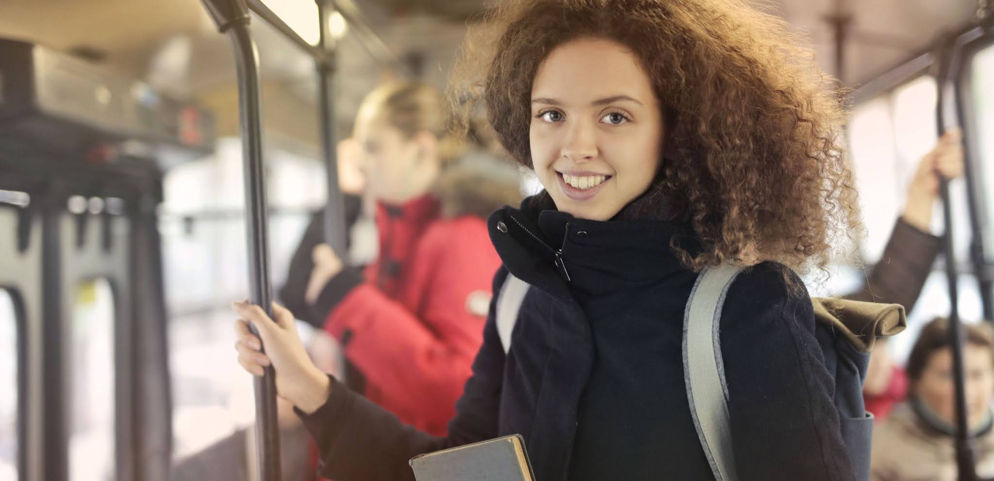 Smiling person holding a pole and a book while riding a city bus with other passengers
