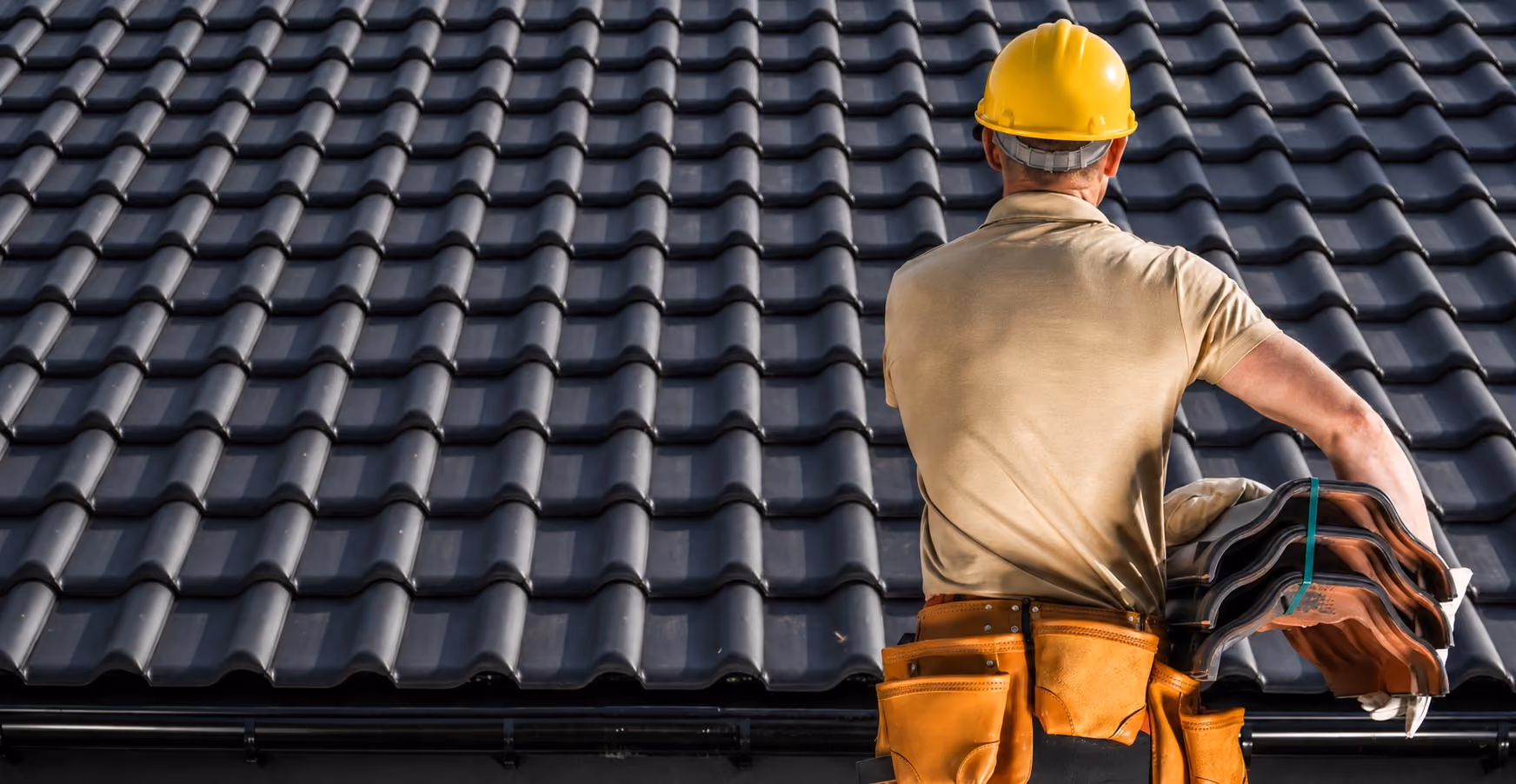 Roofer in yellow hard hat holding roof tiles in front of a newly tiled roof.