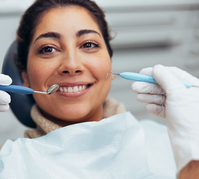 Woman sitting at a dentist chair smiling. There are two hands with dental instruments doing the checkup. 