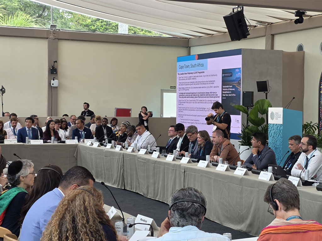 A wide view of an international panel discussion at COP30 in Belém, Brazil. Delegates from around the world are seated at a long conference table beneath a canopy, with a large presentation screen behind them highlighting climate adaptation programs from Cape Town, South Africa. The scene captures active dialogue, global collaboration, and the exchange of ideas on urban sustainability and climate resilience.