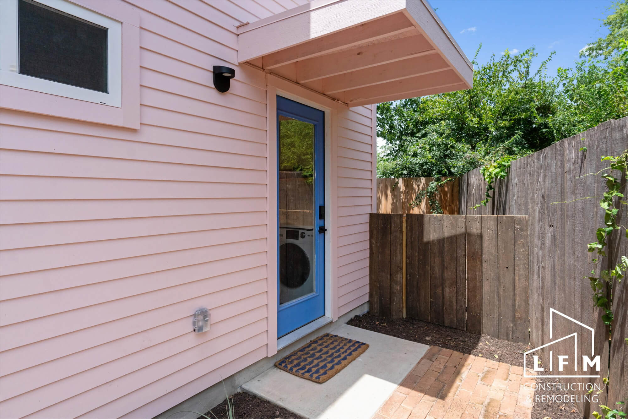 The image shows the entrance to a light pink house with horizontal siding. The door is a vibrant blue with a glass panel, and there is a small welcome mat with a patterned design placed on a concrete step. To the left, there is a window covered with a screen. A light fixture is mounted beside the door. Next to the entrance, there are wooden fence panels that create a private outdoor area, with greenery visible beyond the fence. The ground features a mix of concrete and compacted earth, with a few small plants near the edge.