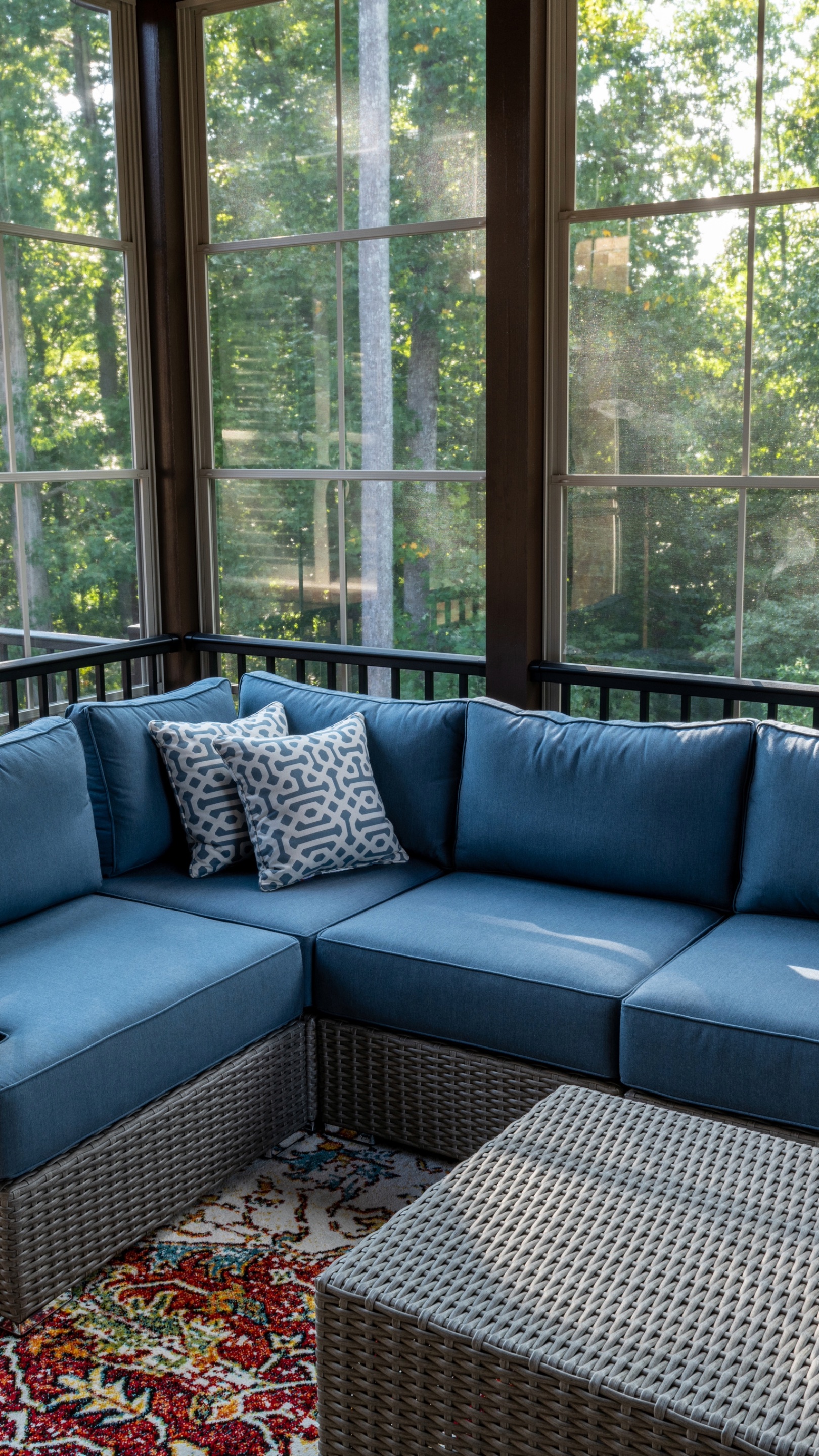 Blue outdoor sectional sofa with patterned pillows in a sunlit room, featuring large windows and lush greenery.