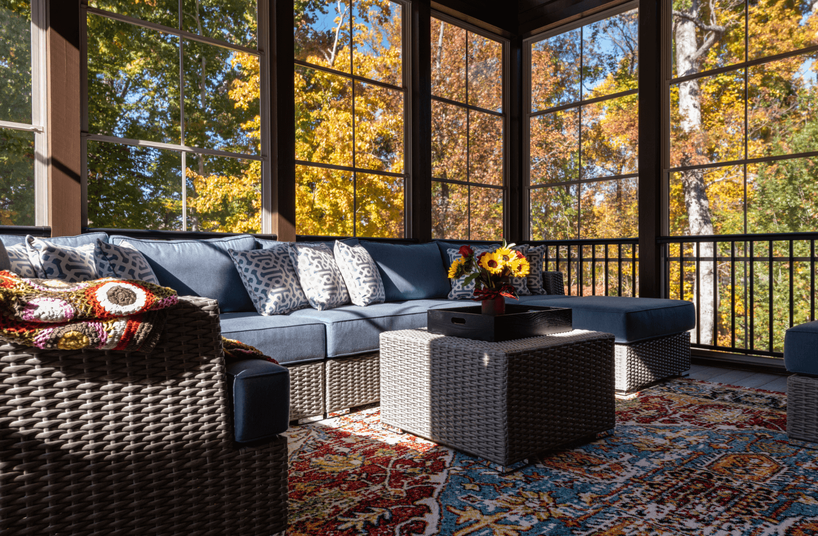 Sunlit screened porch with blue sectional sofa, patterned pillows, and autumn trees visible through windows.