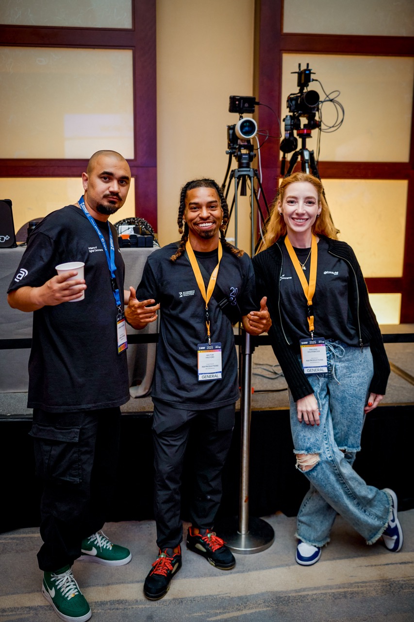 Three production crew members smiling beside video cameras on tripods at a Miami conference