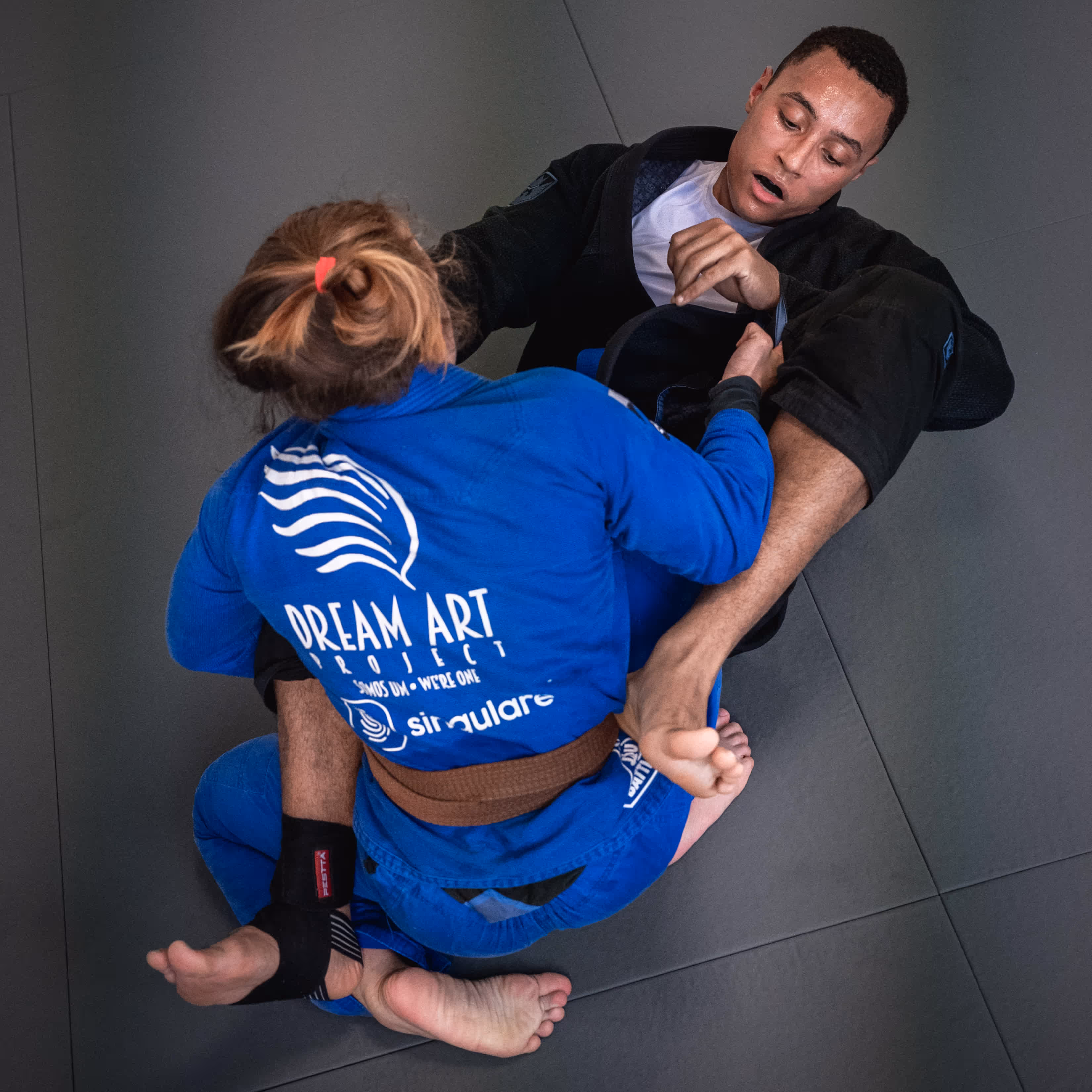 Eine weibliche BJJ-Kämpferin in blauem Gi kontrolliert ihren Trainingspartner mit einer Beinhebeltechnik.